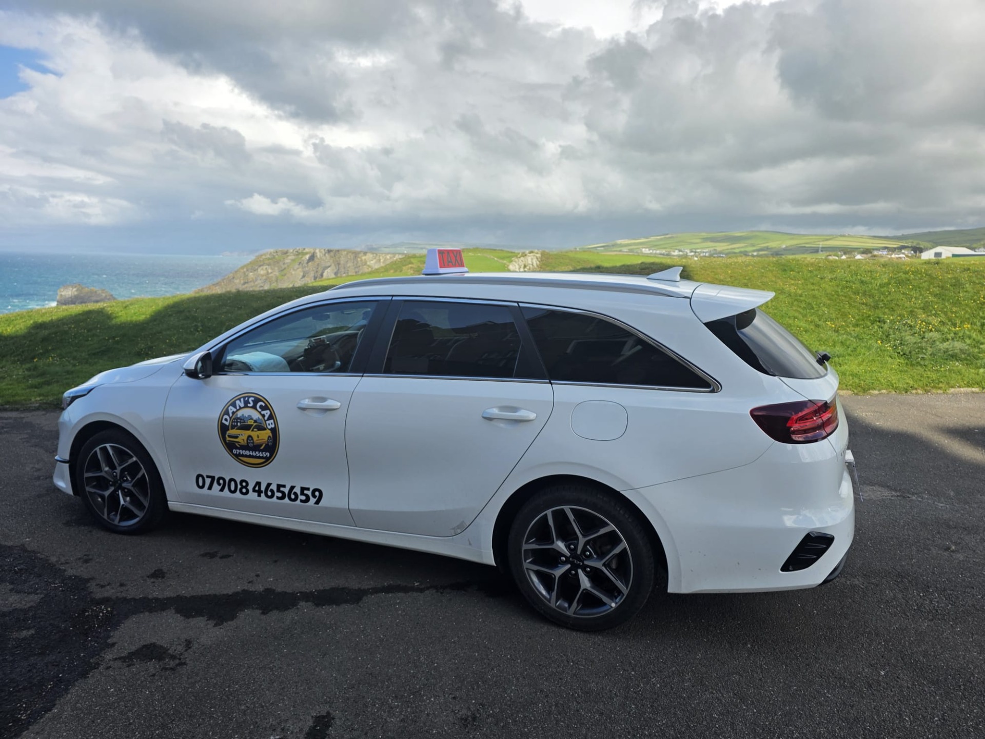 White taxi car parked on coastal road, grassy hills and ocean under cloudy sky in background.
